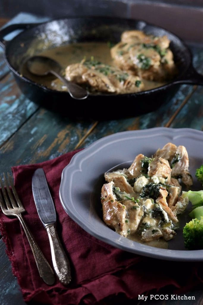 Keto Spinach and Feta Stuffed Chicken Breast with Creamy Sauce - My PCOS Kitchen - A cast iron skillet in the background with stuffed chicken breasts. A grey plate over a burgundy napkin with a chicken breast and a side of broccoli.