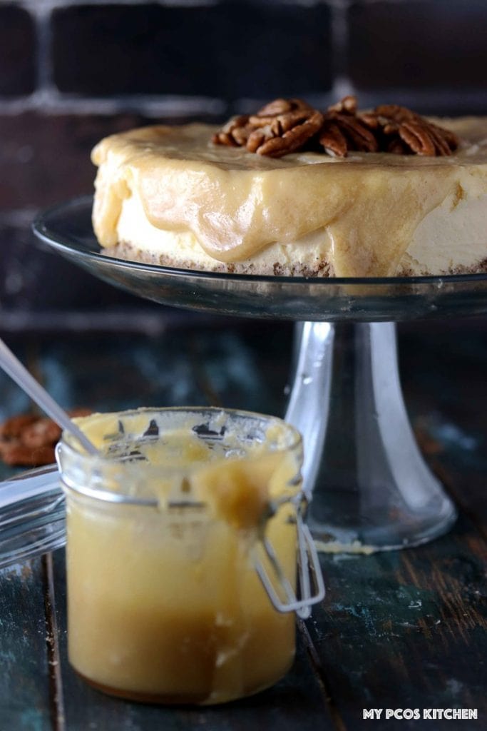 Sugar Free Cheesecake with Caramel - My PCOS Kitchen - Glass of caramel sauce in front of glass cake stand with a gluten-free low carb caramel cheesecake over it.