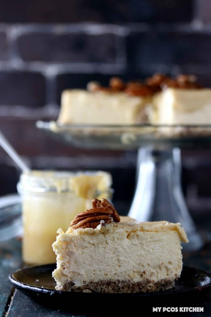Sugar Free Cheesecake with Caramel - My PCOS Kitchen - A slice of sugar-free cheesecake with caramel sauce over. A whole cheesecake on a glass cake stand and caramel sauce in a glass jar in the background.