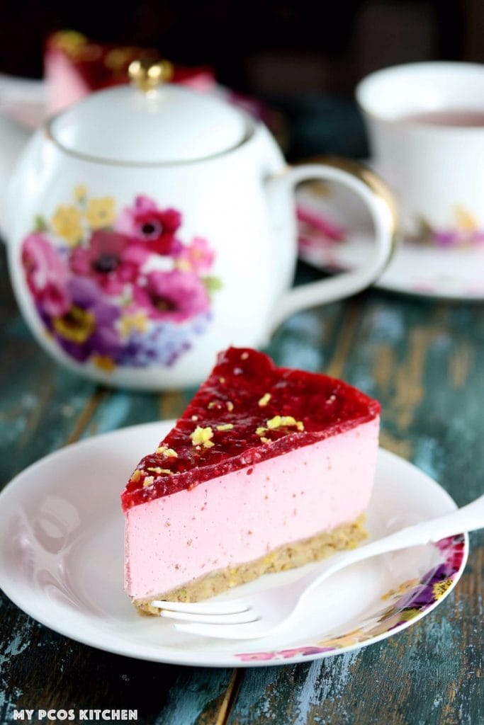 My PCOS Kitchen - Low Carb No Bake Raspberry Lemon Cheesecake (Gluten-free & Sugar-free) - A slice of cheesecake cut over a flower plate with a white fork. Beautiful flower tea kettle behind cheesecake.