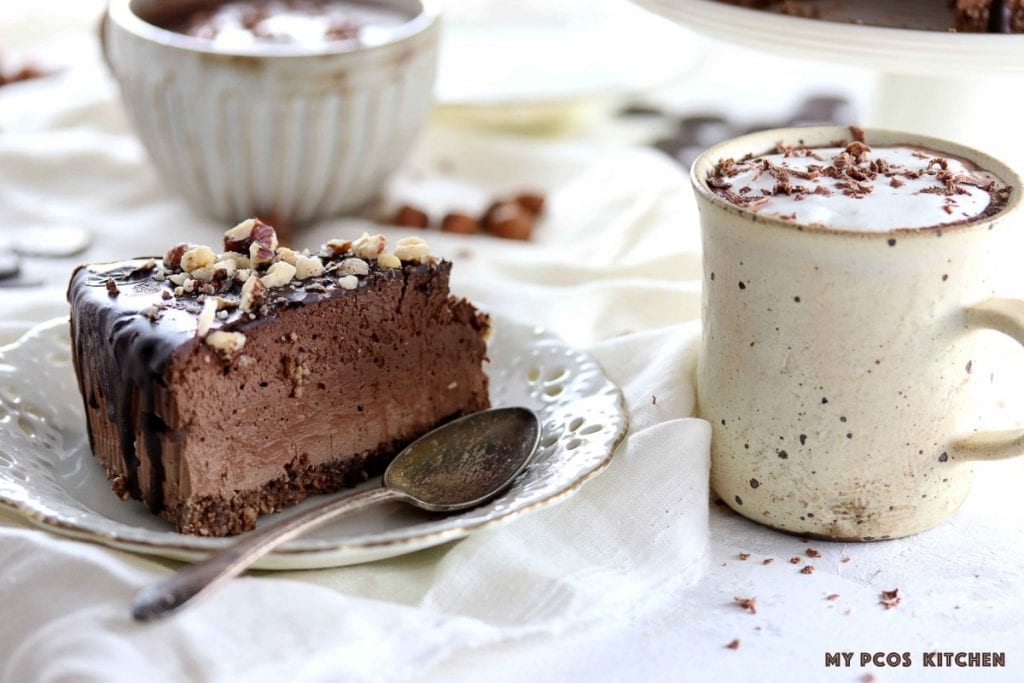 Low Carb Keto Triple Chocolate Cheesecake - My PCOS Kitchen - A thick slice of chocolate cheesecake over a white floral plate. A vintage silver spoon on the side and two handmade ceramic cups filled with hot chocolate.