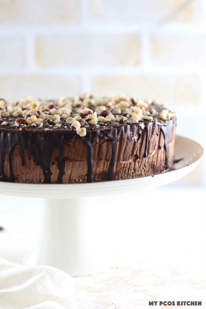Low Carb Keto Triple Chocolate Cheesecake - My PCOS Kitchen - A whole chocolate cheesecake with a chocolate ganache drizzling from the sides with crushed hazelnuts on the top of the cake. Over a white cake stand from H&M Home.