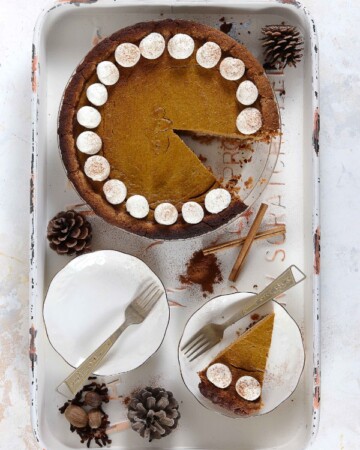 Photo of pumpkin pie with a slice cut on a white plate over a white metal tray.
