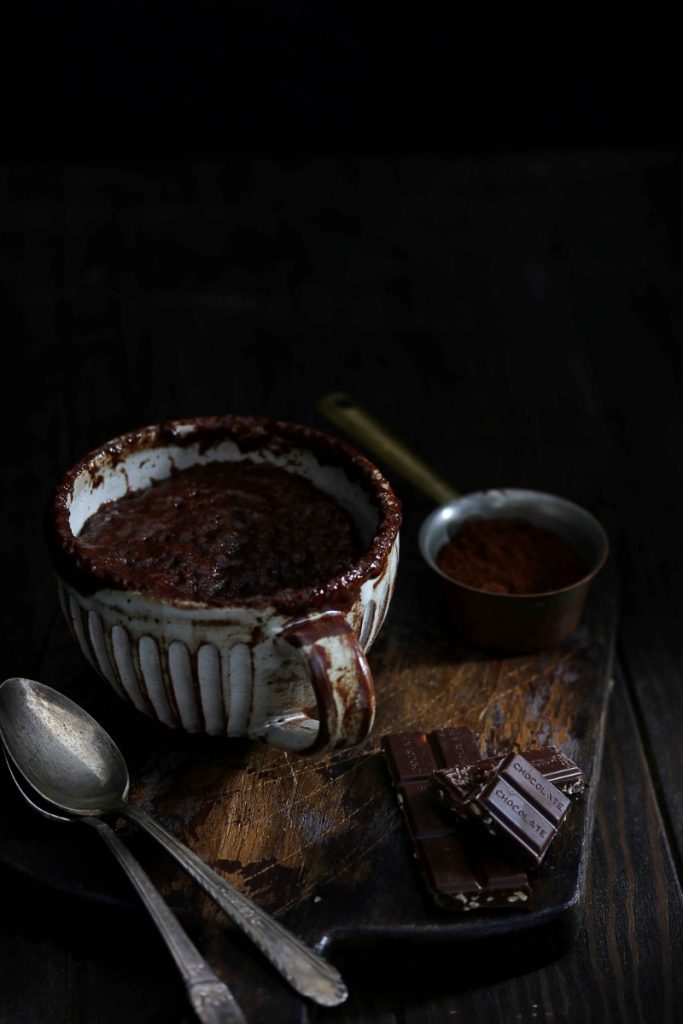 A keto chocolate mug cake on a wooden board beside some chocolate and espresso powder.