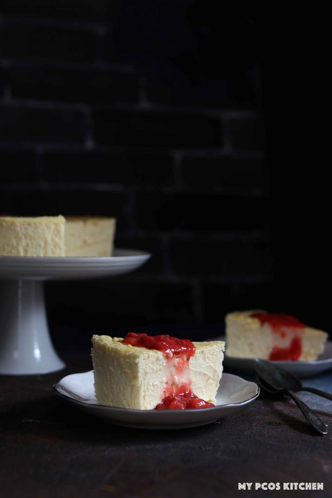 Two slices of sugar free cheesecake with a low carb strawberry sauce poured over on two small plates with a whole cheesecake in the background on a white cake stand.