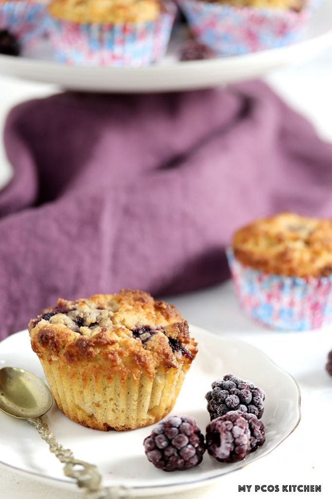 A closeup shot of a keto blackberry muffin on a white plate beside frozen blackberries.